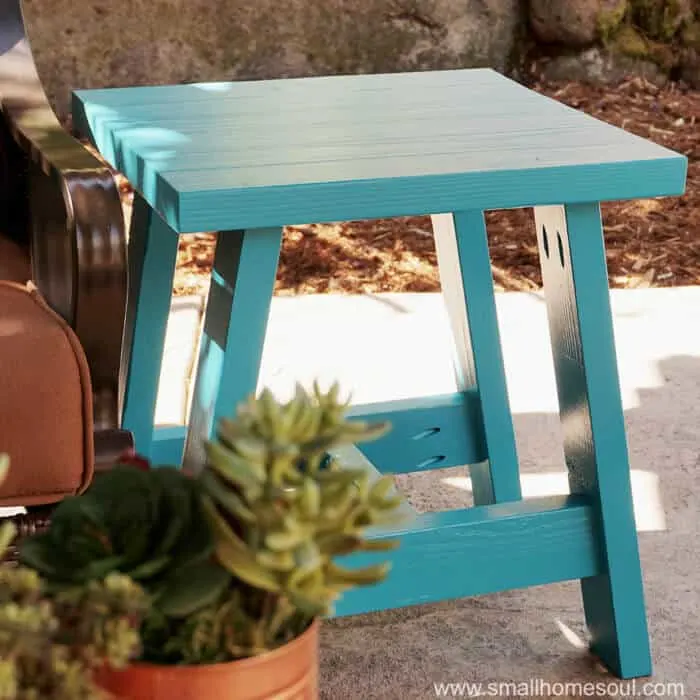 Turquoise-painted outdoor side table with a slatted top and angled legs sits beside a patio chair.