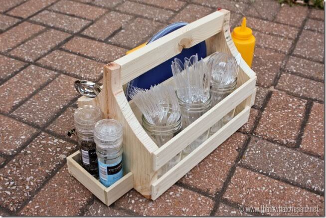 Wooden tailgating caddy holding plastic utensils, plates, cups, condiments, and shakers on a patio.