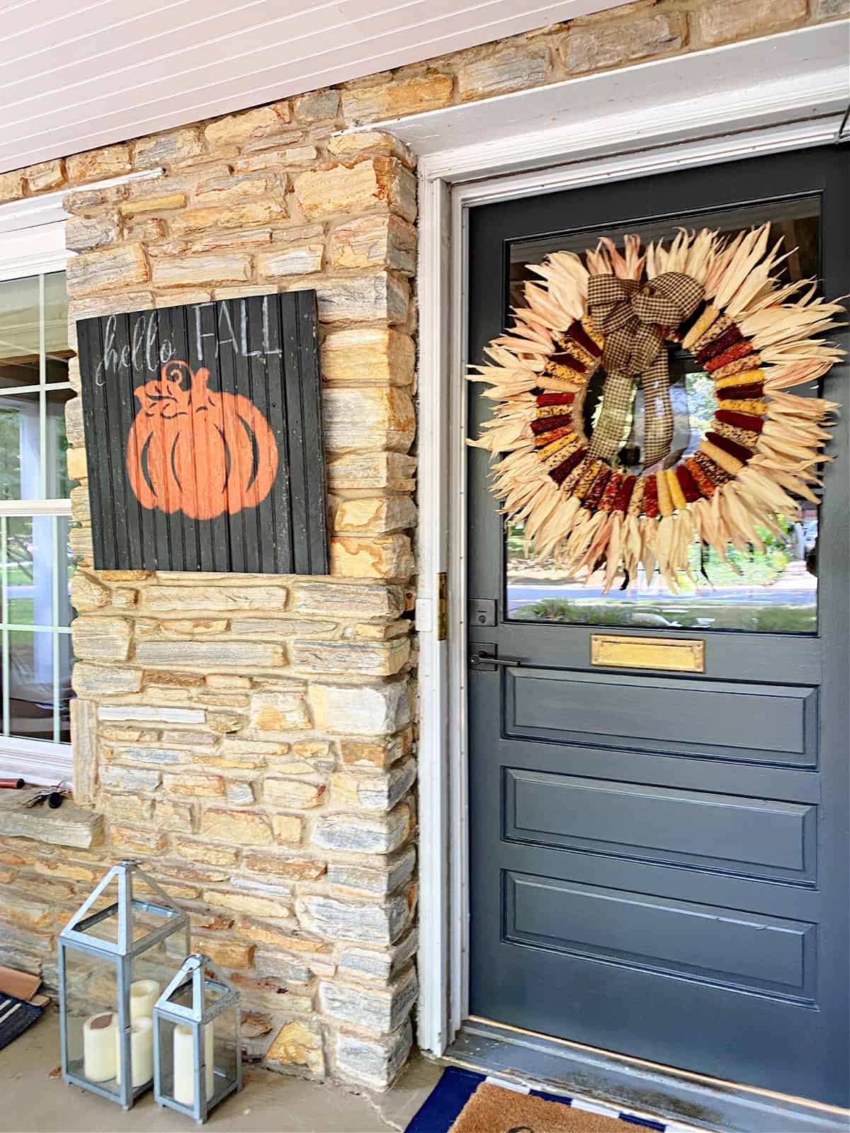 Black painted sign with an orange pumpkin that says, "Hello Fall" on a brick wall.