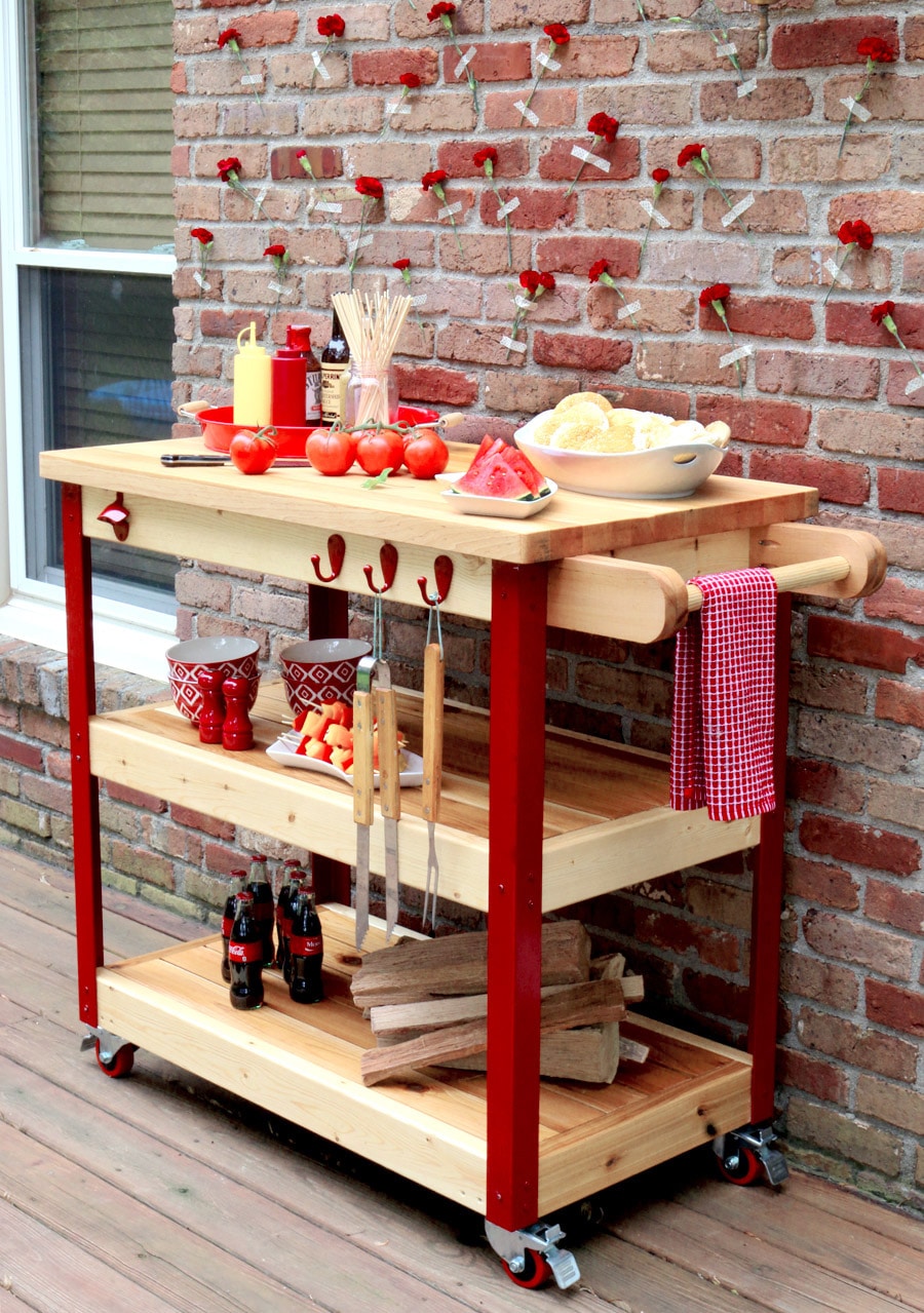 DIY outdoor grill cart with a butcher block-style top, two lower storage shelves, hanging grilling tools, towel bar, locking casters, and red painted legs on a backyard deck against a brick wall decorated with red flowers.