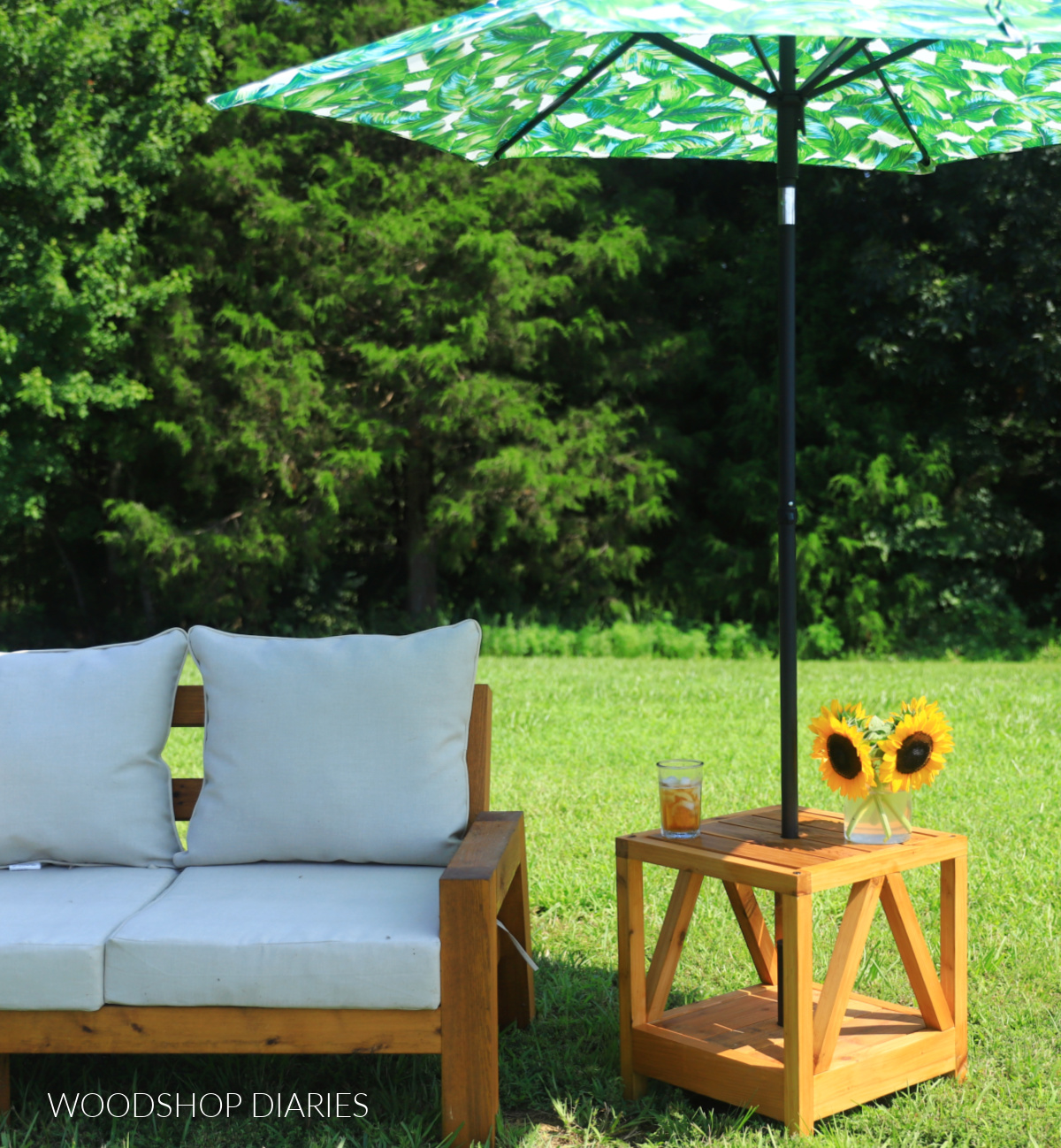 DIY outdoor side table with a built-in umbrella stand sits beside a cushioned patio sofa on a grassy lawn.
