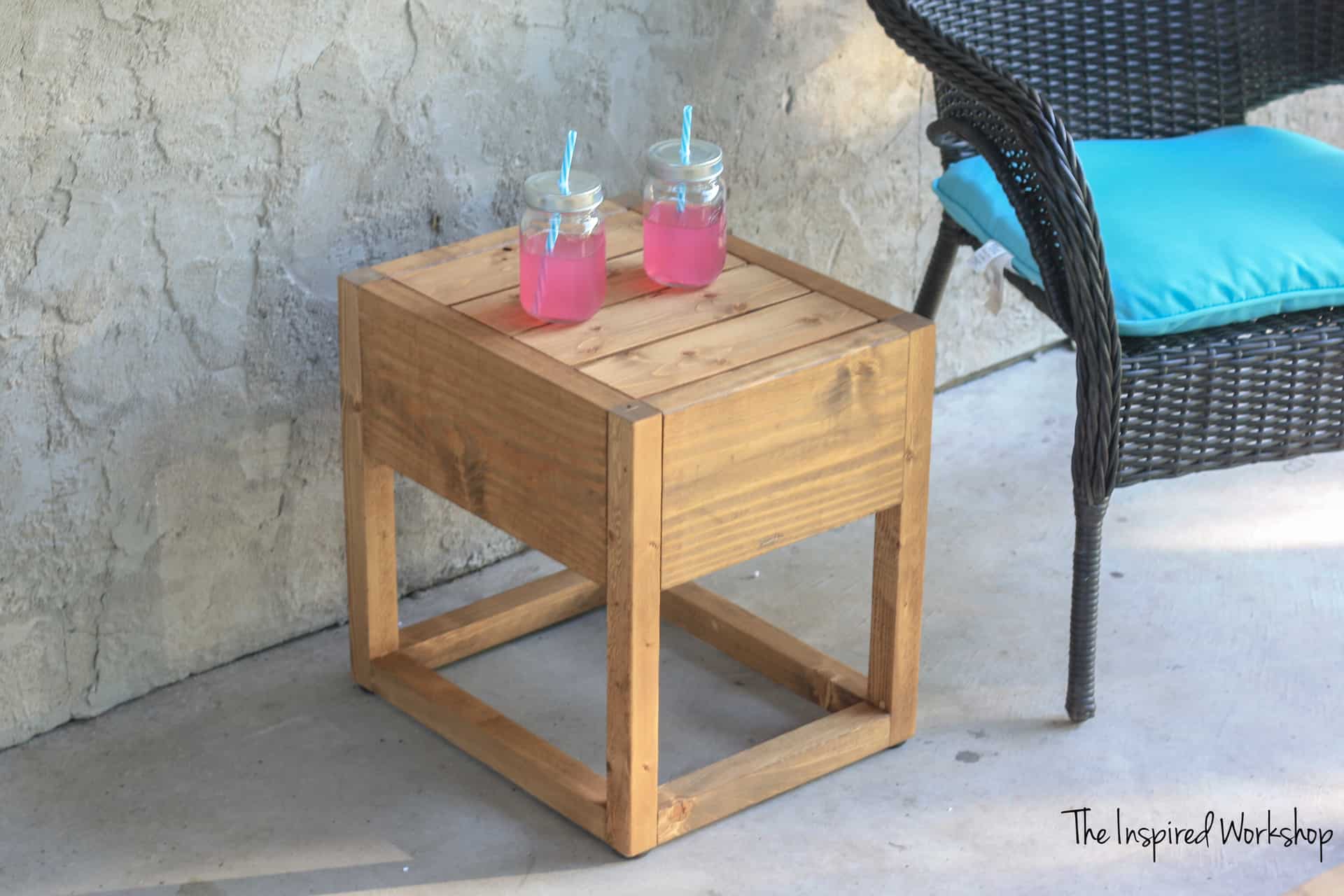 DIY outdoor side table with a slatted wood top and open base sits beside a wicker patio chair.