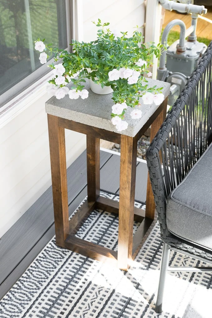 Tall outdoor side table with a concrete top and stained wood base stands beside a patio chair.