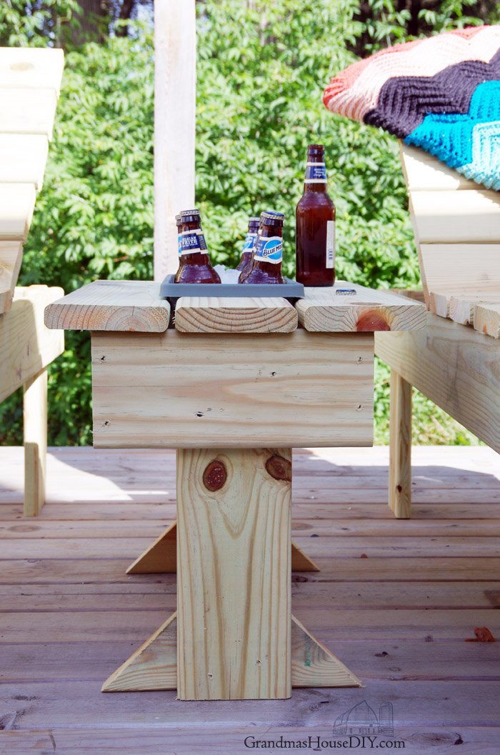 DIY outdoor side table with a built-in cooler compartment sits between two Adirondack chairs on a deck.