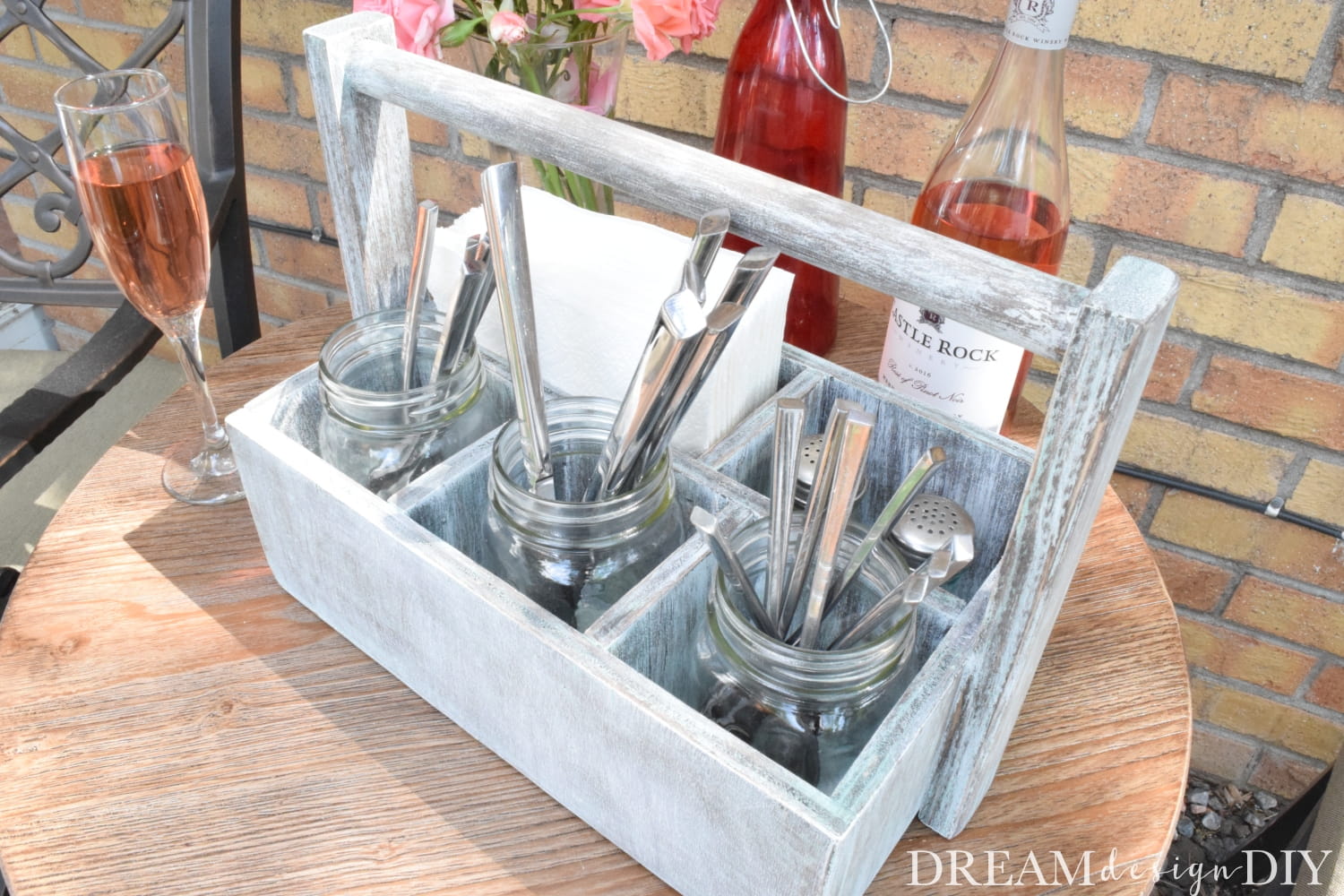 Whitewashed wooden utensil caddy with mason jars holding flatware, napkins, and wine bottles on an outdoor table.