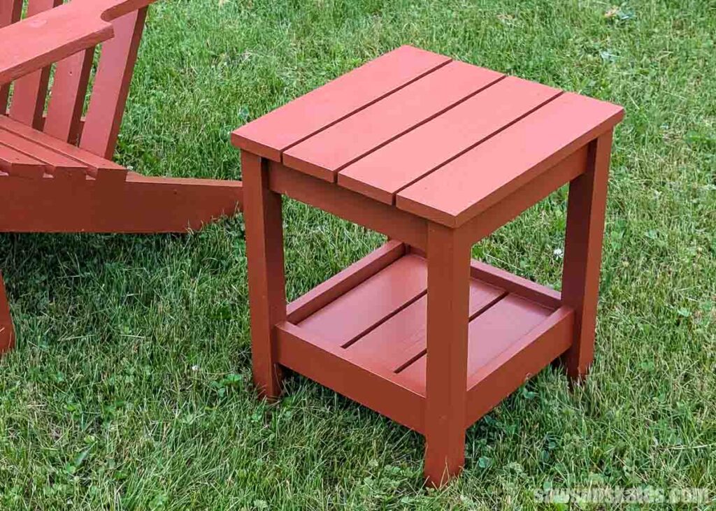 Small DIY outdoor side table with a lower storage shelf, painted reddish brown and placed on a lawn beside a matching Adirondack chair.
