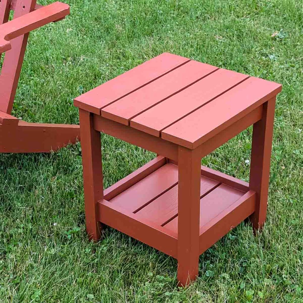 Small DIY outdoor side table with a lower storage shelf, painted reddish brown and placed on a lawn beside a matching Adirondack chair.
