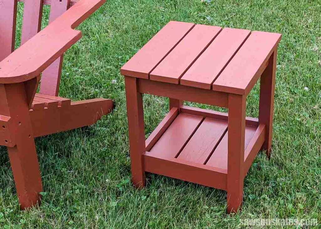 DIY outdoor side table with a slatted top and lower shelf, painted a warm reddish-brown and positioned on a grassy lawn beside an Adirondack chair.