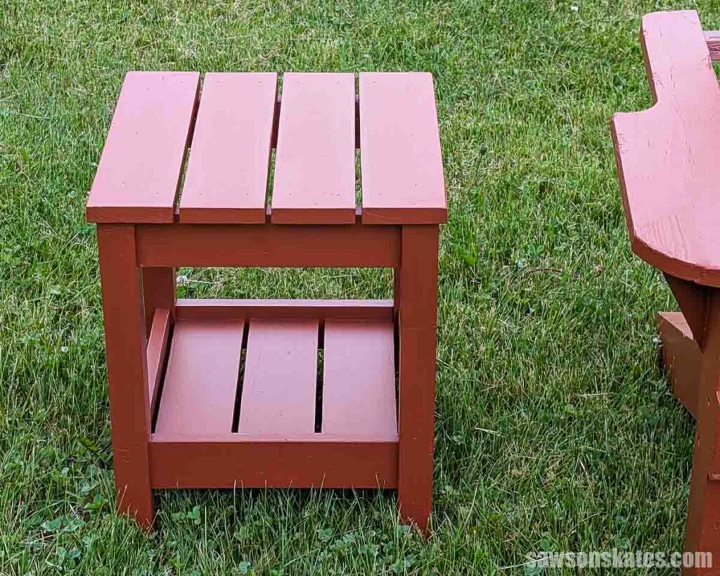Front view of a DIY outdoor side table painted reddish brown, featuring a slatted square top, straight legs, and a lower slatted storage shelf.