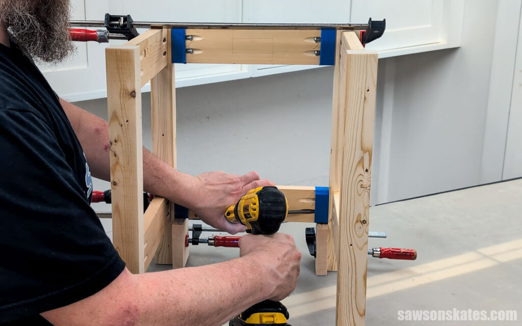 Woodworker attaching the lower shelf supports to a DIY outdoor side table frame using pocket hole screws and a cordless drill. Bar clamps and spacer blocks hold the parts in position while the table base is assembled on a workshop bench.