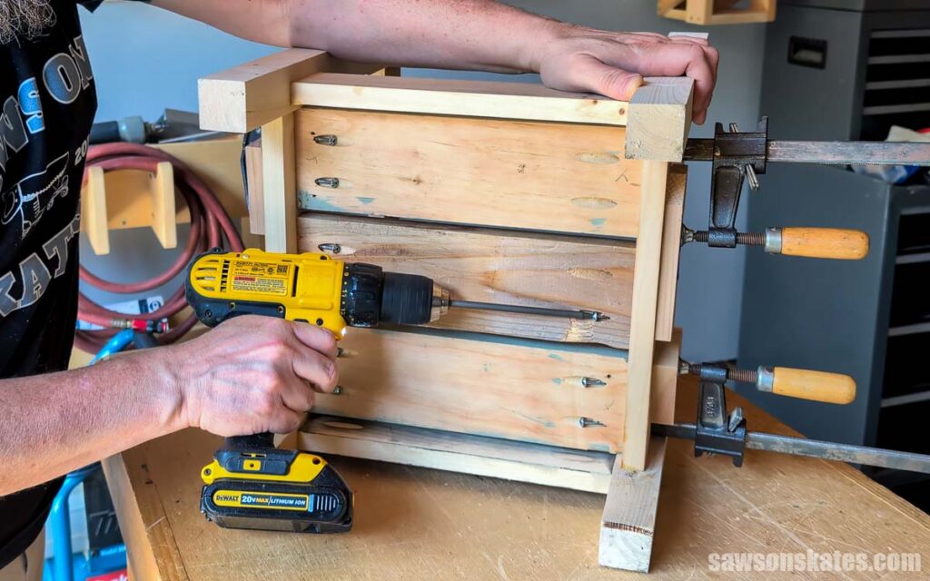 Person using a cordless drill to attach the slatted lower shelf to a wooden side table frame. Bar clamps hold the assembly in place while pocket hole screws are driven into the shelf boards from underneath.