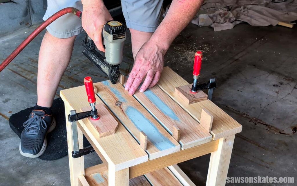 Person using a brad nailer to attach slats to the top of a wooden side table. Clamps and spacer blocks hold the boards evenly spaced while the slats are fastened to the table frame.