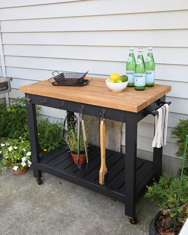 Black DIY outdoor grill cart with a butcher block-style top, lower slatted shelf, side towel bar, hanging hooks for grilling tools, and locking casters on a backyard patio.