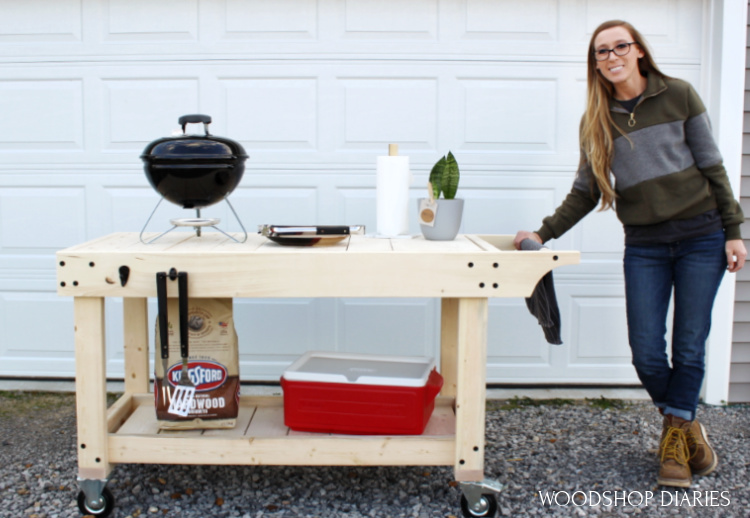 DIY outdoor grill cart with a large wood top, lower storage shelf, locking casters, and a charcoal grill on top, with a person standing beside it in a driveway.