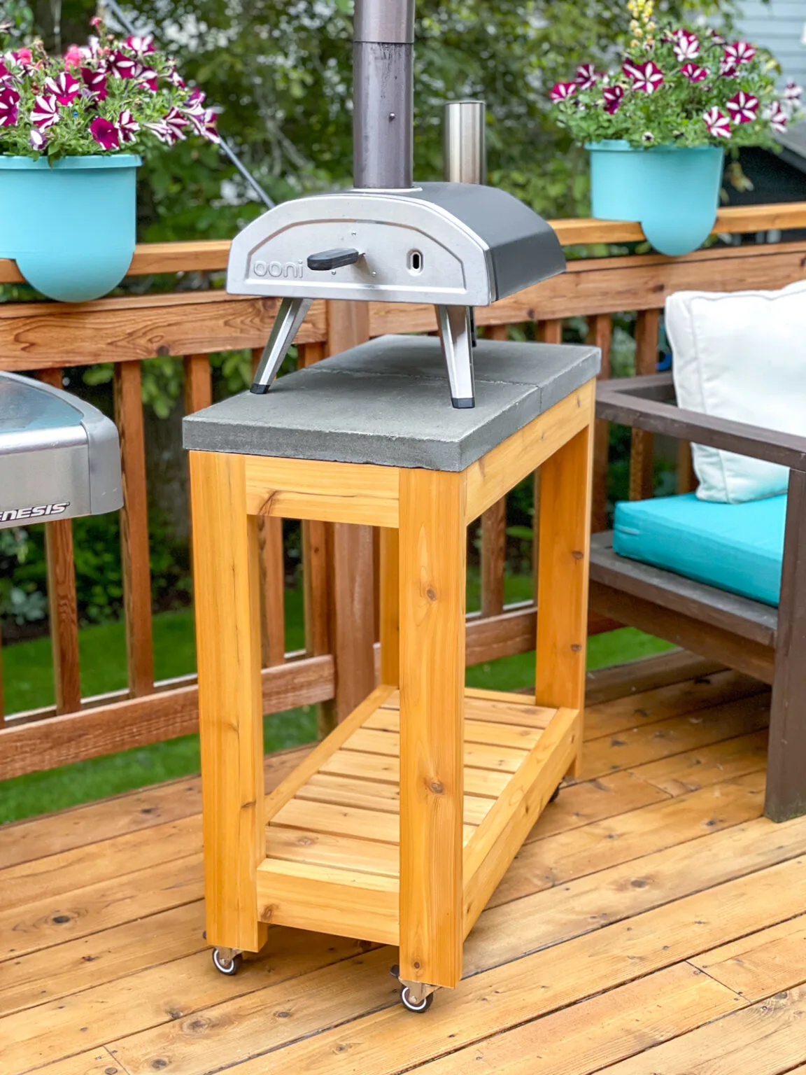 Outdoor pizza oven sitting on a rolling cedar stand with a concrete top and lower slatted shelf on a backyard deck with flower planters and patio furniture in the background.