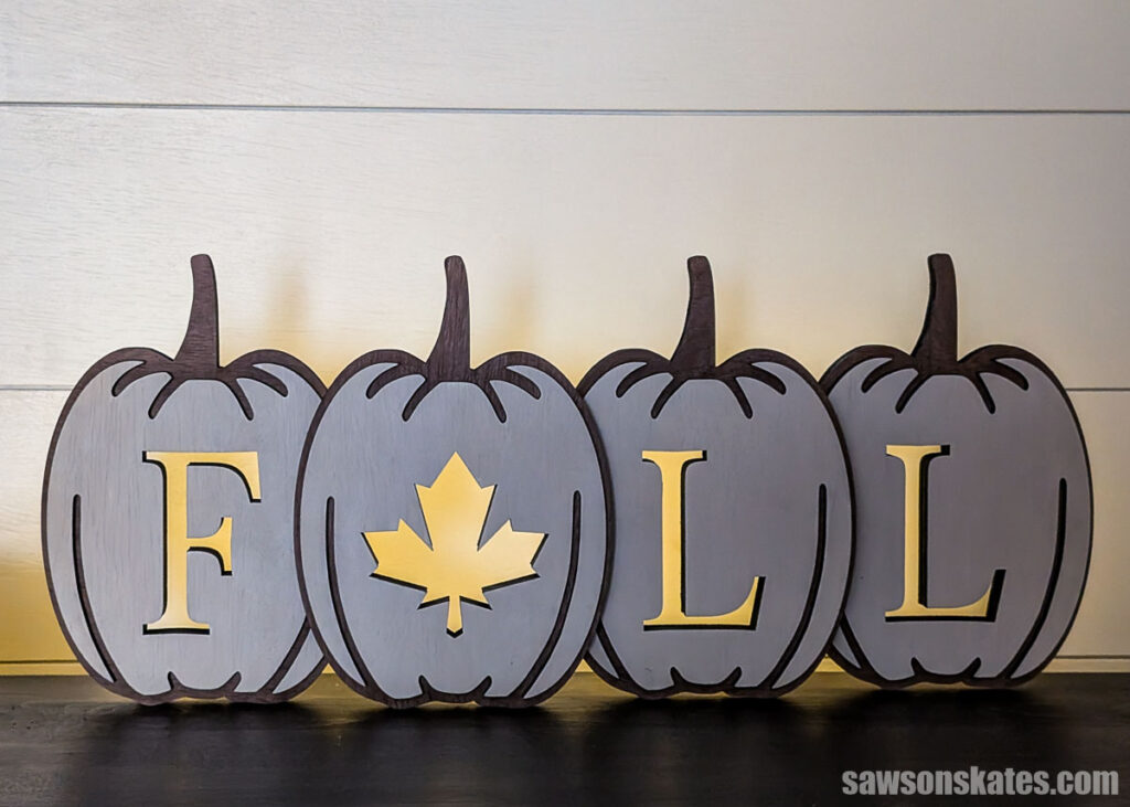 Front view of a layered pumpkin fall sign in a dark room with lights behind the sign