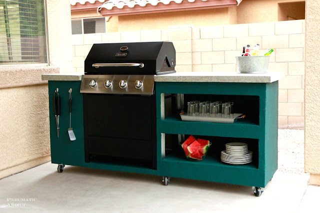 DIY outdoor grilling station with a built-in gas grill, open storage shelves, side tool hooks, locking casters, and a concrete-style countertop on a backyard patio.