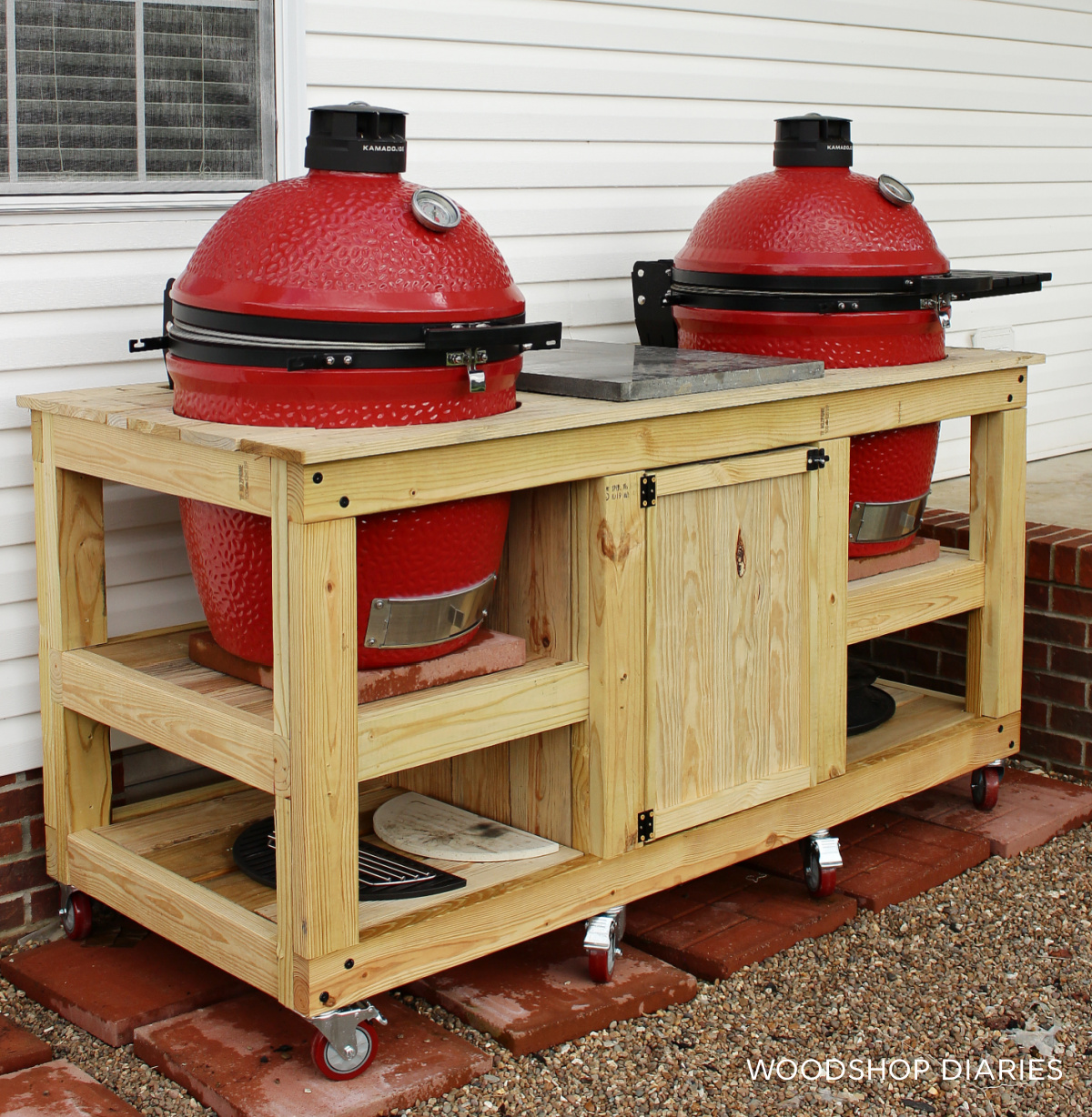 DIY outdoor grilling station with two red kamado-style grills built into a large rolling wood cart with storage shelves, cabinet doors, and locking casters beside a house.