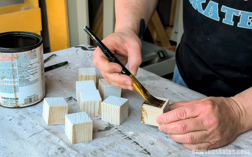 Close-up of hands applying dark stain with a paintbrush to small unfinished wood blocks on a workbench.