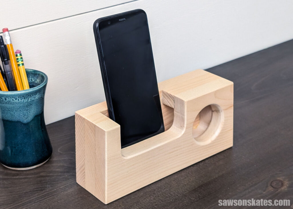 Wooden passive speaker holding a smartphone in a curved slot with a round speaker opening, placed on a desk next to a cup of pencils.