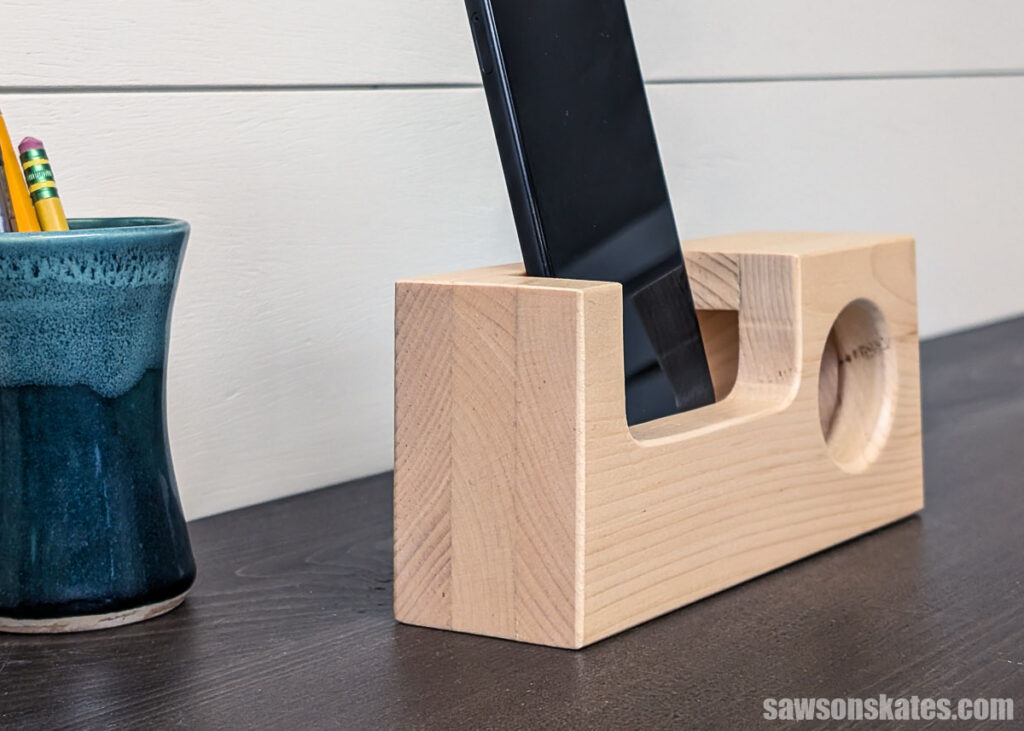 Wooden phone speaker holding a cell phone in a curved slot, with a round speaker opening, on a desk next to a cup with pencils.
