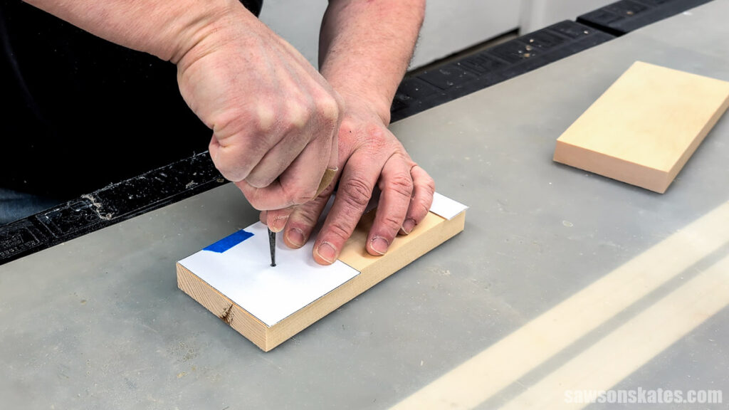 Close-up of hands marking a small wood block with an awl through a paper template