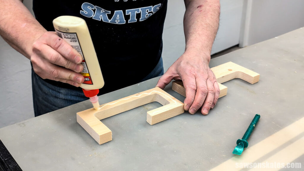 Applying wood glue along the inside edge of a U-shaped wooden piece on a work surface, with a second piece nearby.