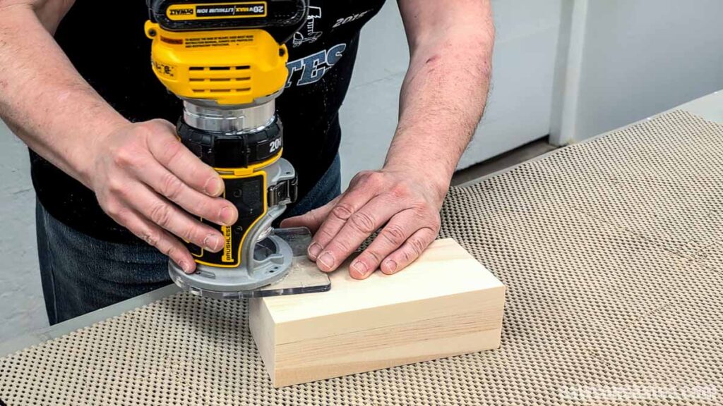 Hands using a trim router to round over the edges of a rectangular wood block on a non-slip mat.