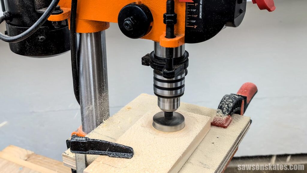 Drill press cutting a large hole in a wood block with a Forstner bit, the piece secured with clamps and covered in sawdust.