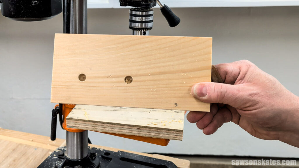 Hand holding a wood block with two evenly spaced drilled holes in front of a drill press