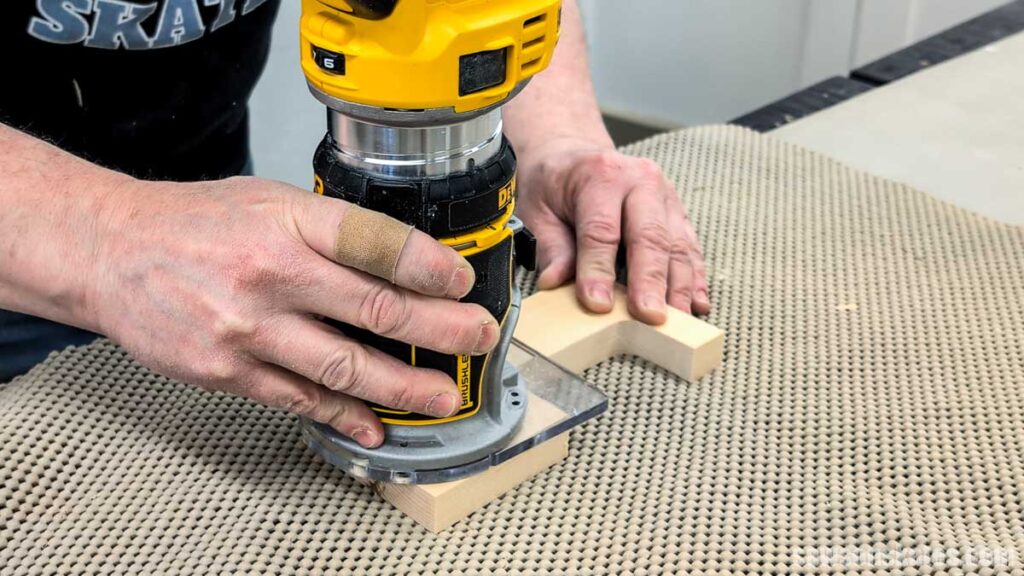 Hands using a trim router to round over the edges of a wooden piece on a non-slip mat.