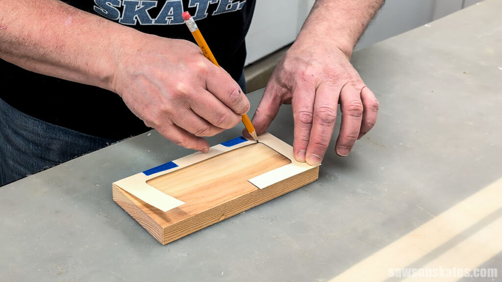 Hands using a pencil to trace a rectangular opening onto a wood block with a taped template on a workbench.