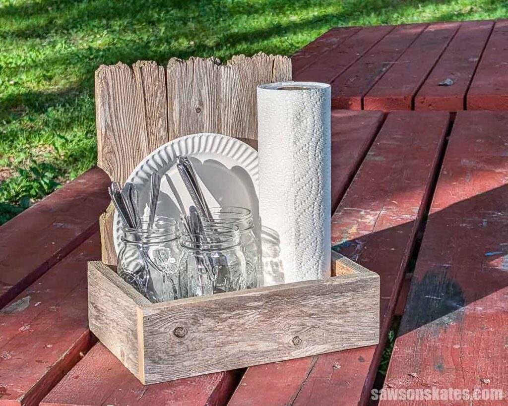 Rustic wooden picnic caddy holding mason jars of utensils, paper plates, and a roll of paper towels on a red picnic table.