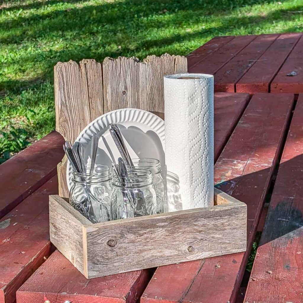 Rustic wooden picnic caddy holding mason jars of utensils, paper plates, and a roll of paper towels on a red picnic table.