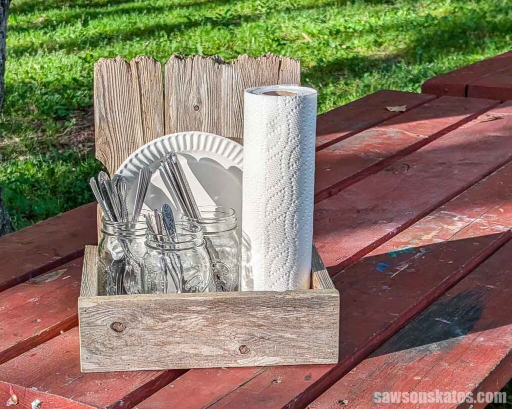 Front view of a wooden picnic caddy holding mason jars of utensils, paper plates, and a roll of paper towels on a red picnic table.