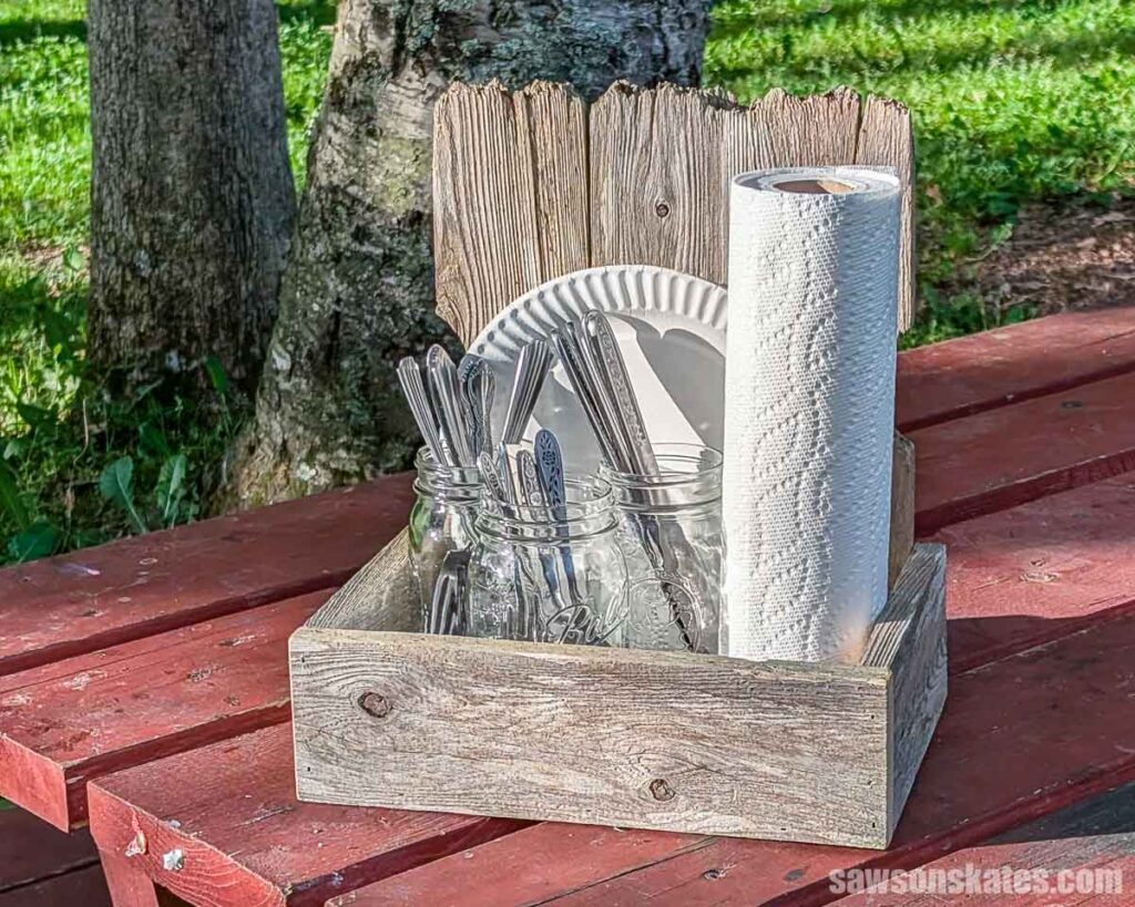 Rustic wooden picnic caddy holding mason jars of utensils, paper plates, and a roll of paper towels on a red picnic table.
