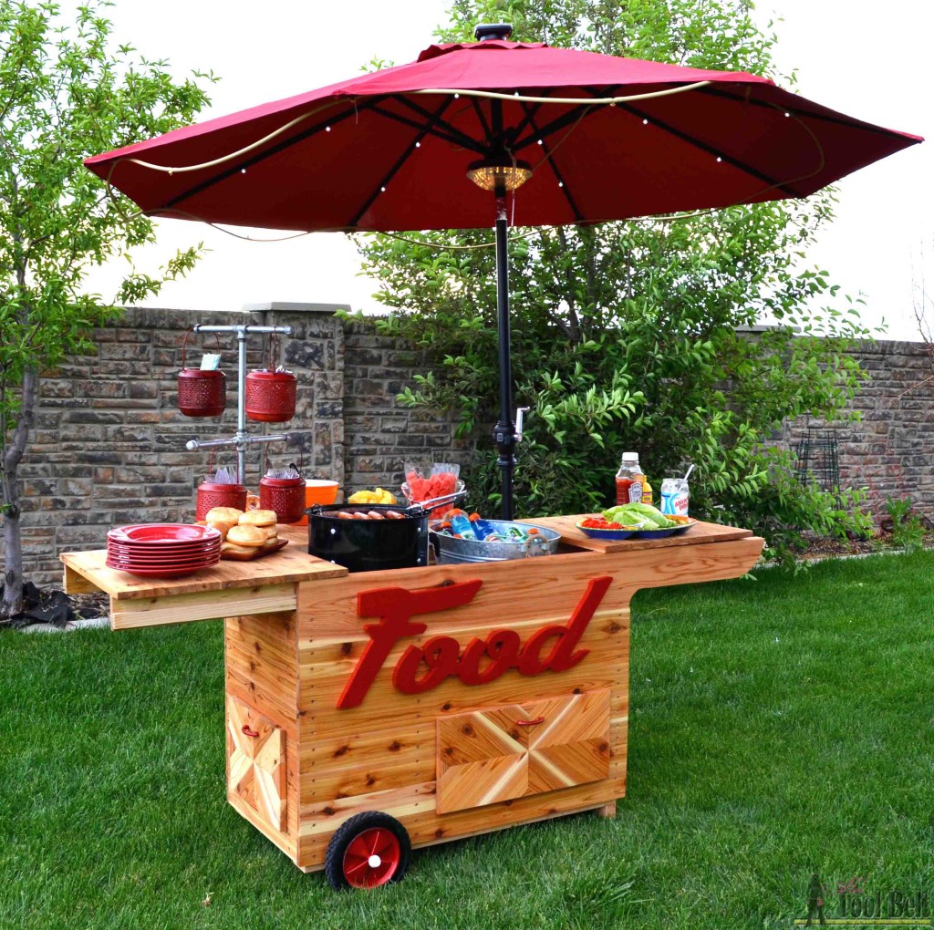 DIY outdoor grill cart with a large patio umbrella, wood serving counter, rolling wheels, storage cabinets, and red “Food” lettering on the front, set up for outdoor entertaining on a backyard lawn.