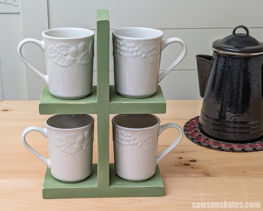 Front view of a DIY stacked mug stand painted green with a distressed finish, holding four white mugs on a wood counter beside a black coffee pot