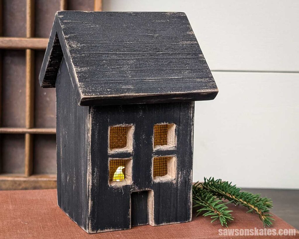 DIY wood house luminary with an LED candle inside sitting on a book with a sprig of evergreen on the right side