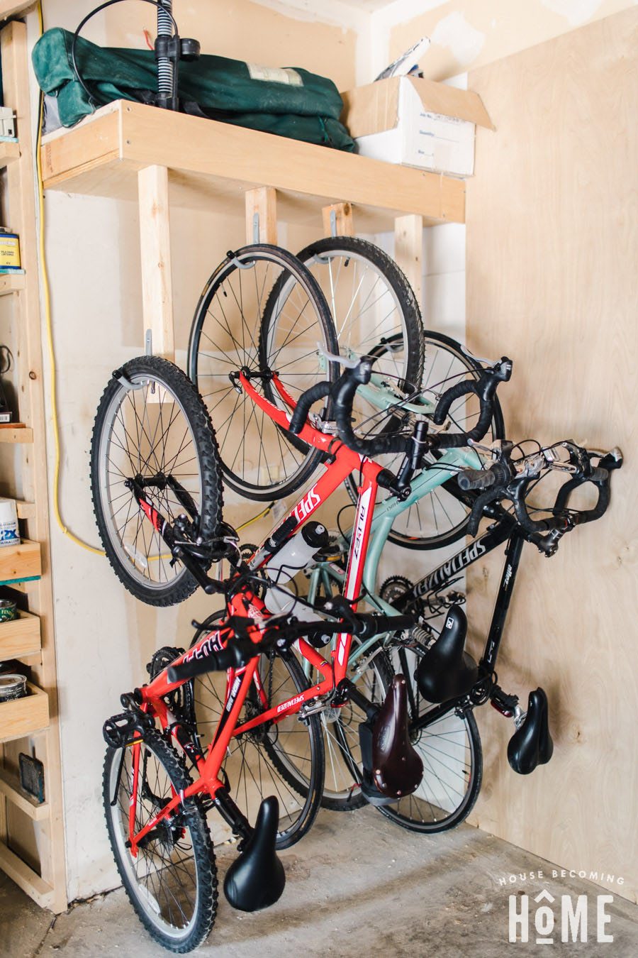 Garage bike storage area with several bicycles hanging vertically from wall hooks below an overhead storage shelf.