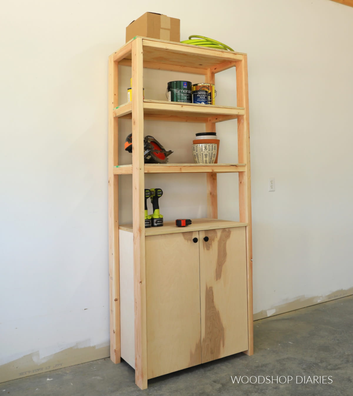 Tall wooden garage storage cabinet with open shelves, double doors, and tools and paint cans stored inside.