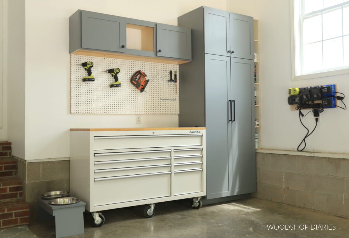 Garage workshop storage area with gray cabinets, a rolling tool chest, pegboard tool storage, and wall-mounted battery chargers.