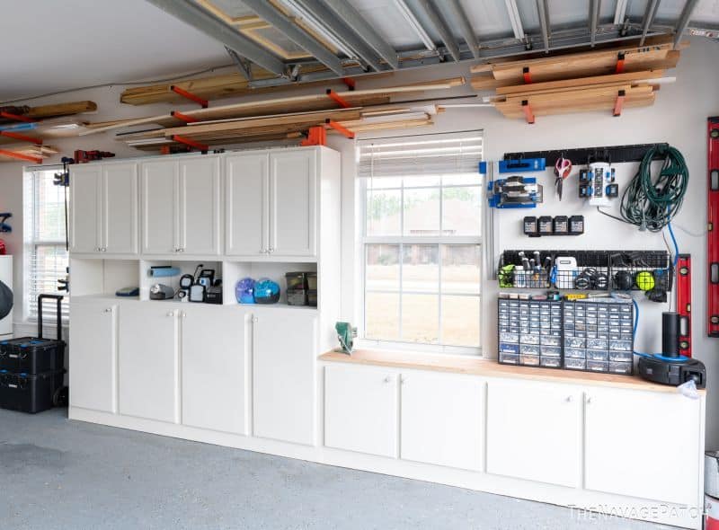 Garage storage wall with white cabinets, a workbench, small parts organizers, wall-mounted tools, and lumber stored overhead.
