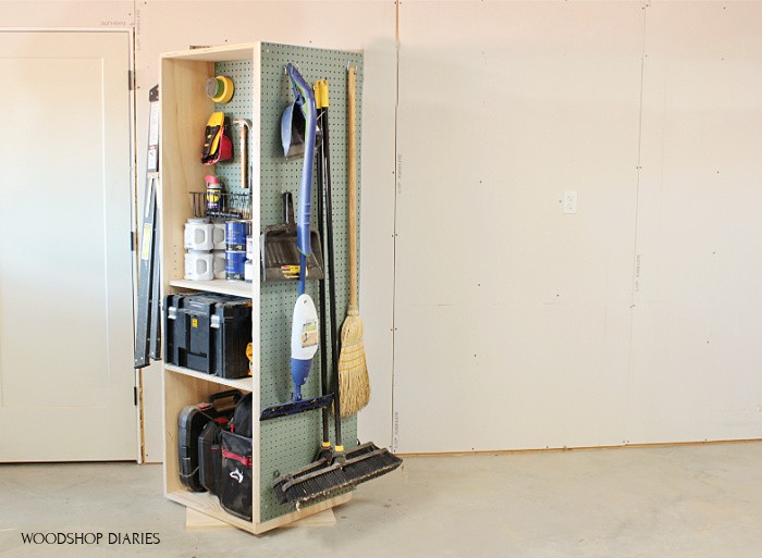 Rotating workshop storage cart with shelves on one side and pegboard holding brooms, mops, and long-handled tools on the other side.