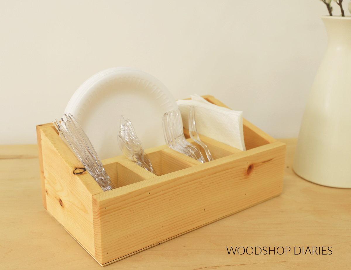 Wooden utensil caddy holding plastic forks, spoons, knives, paper plates, and napkins on a table.