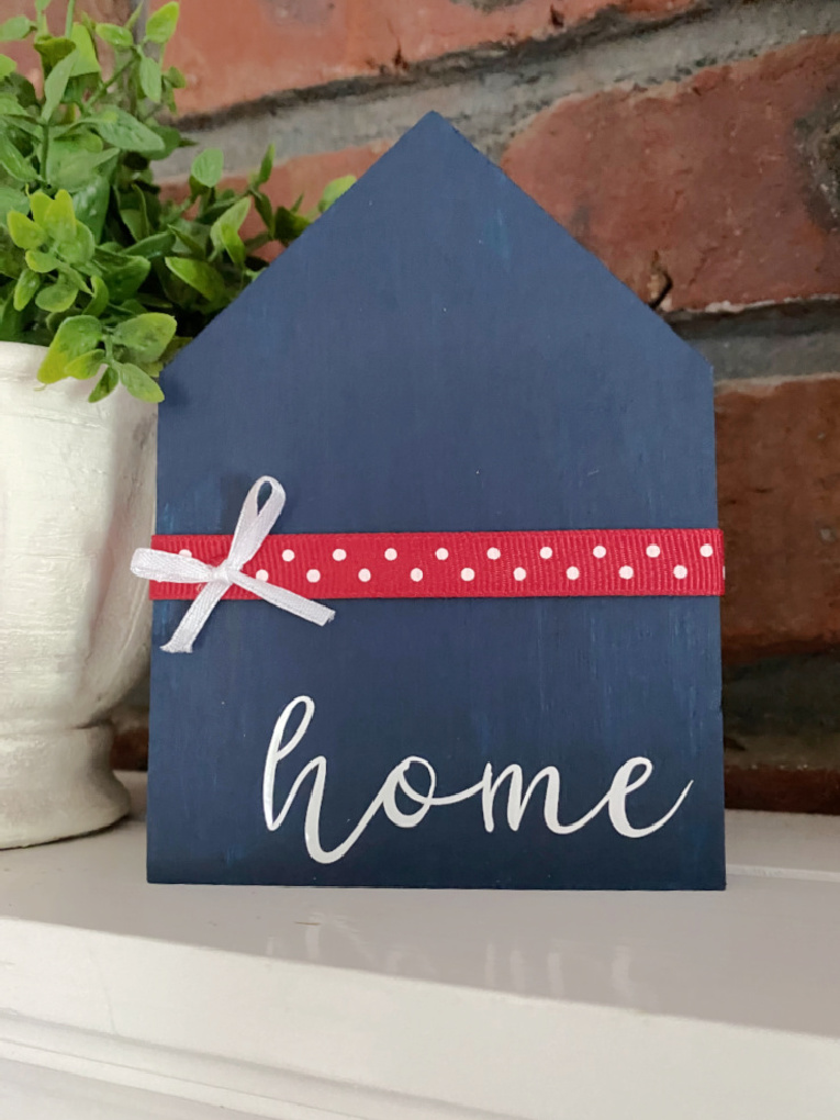 Small house-shaped wooden sign painted dark blue, sitting on a white mantel in front of a brick wall.