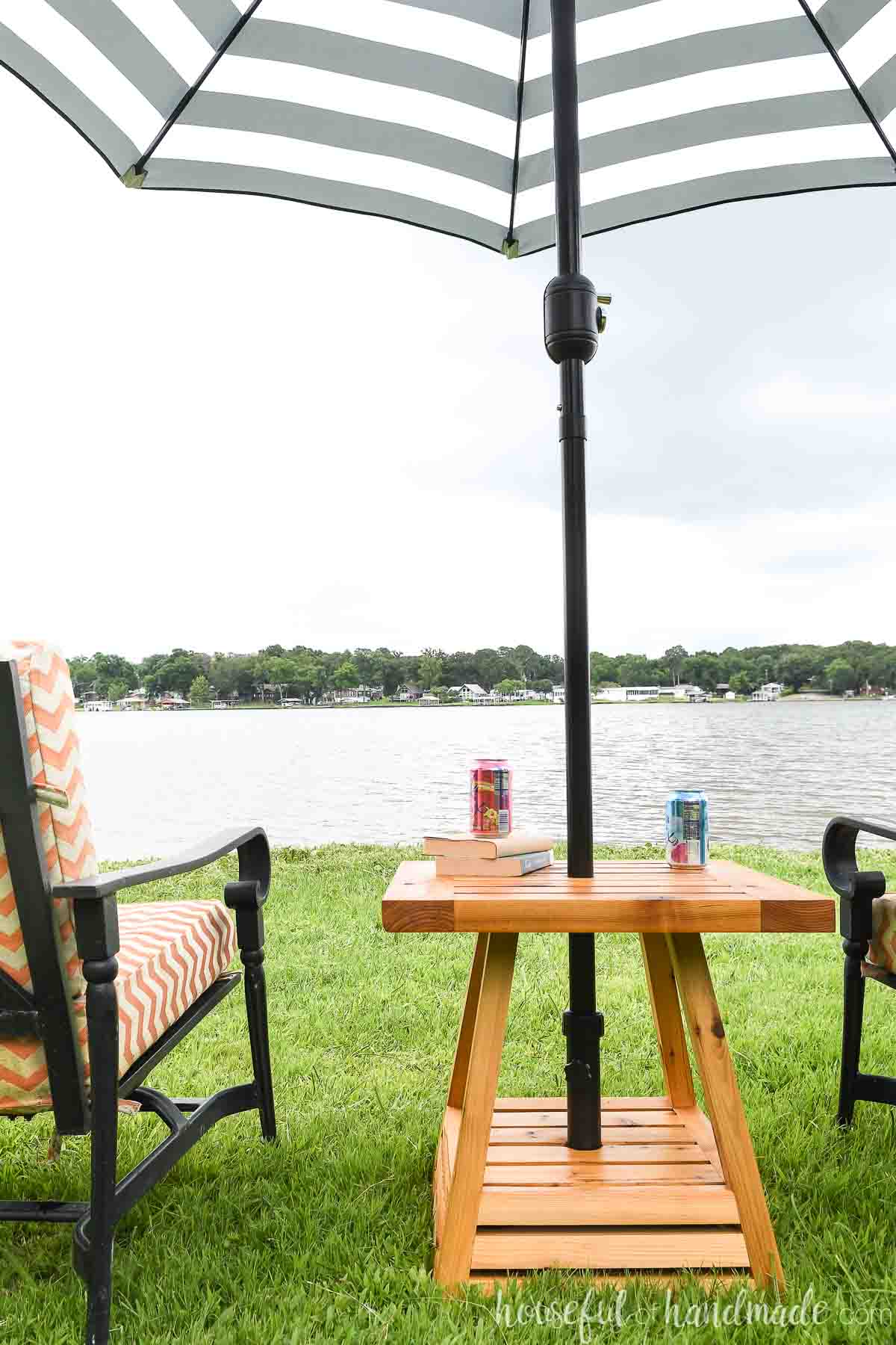 Wooden outdoor side table with a built-in umbrella hole, positioned between two patio chairs overlooking a lake.