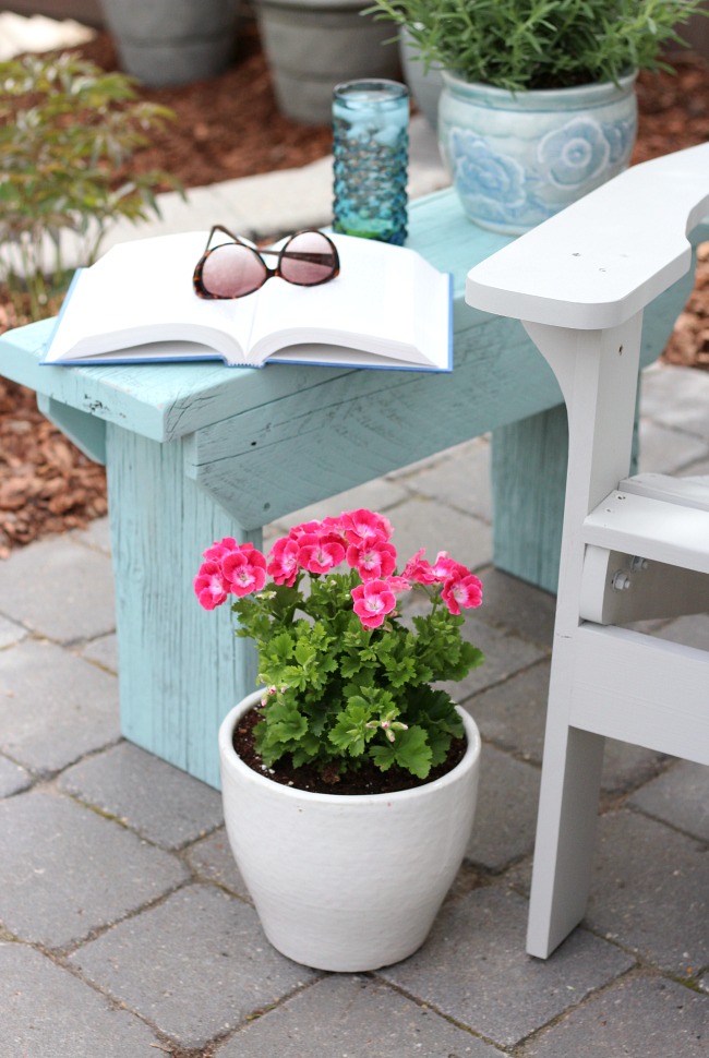 Light blue wooden outdoor side table sits beside a white Adirondack chair on a patio.