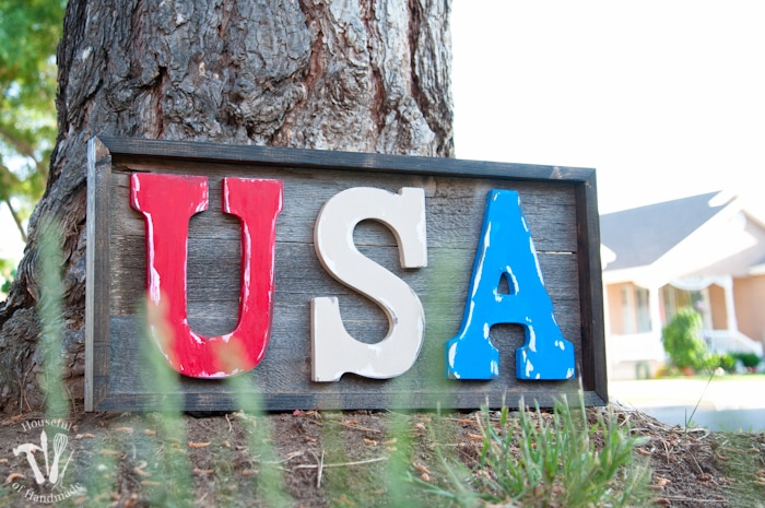 Patriotic wooden sign featuring large red, white, and blue letters spelling ‘USA’ mounted on a rustic wood plank background inside a dark stained frame.