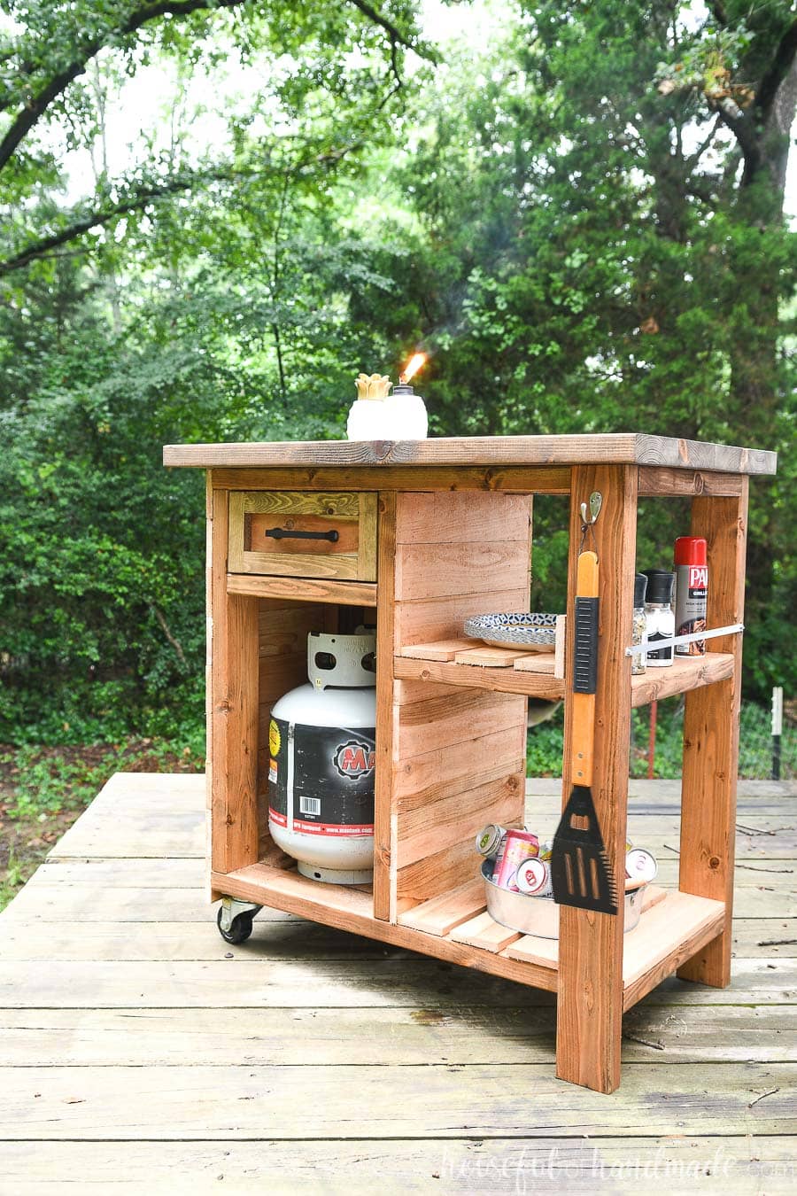 Rustic DIY grill cart with a wood plank top, propane tank storage, small drawer, open shelves, hanging grilling tools, and locking casters on a backyard deck.